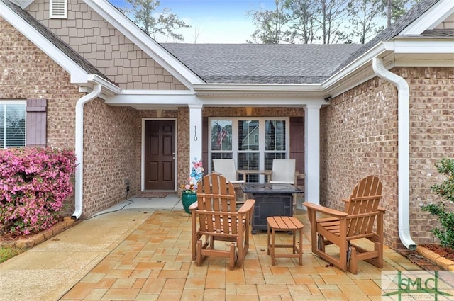 view of exterior entry featuring brick siding, a shingled roof, and a patio area