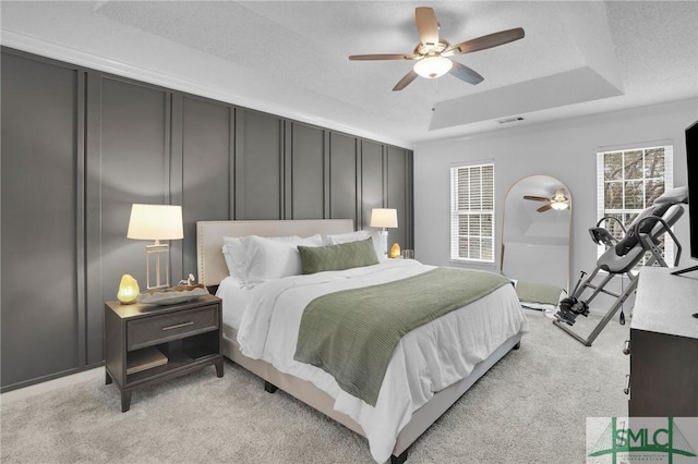 bedroom with visible vents, a ceiling fan, a decorative wall, a raised ceiling, and light colored carpet