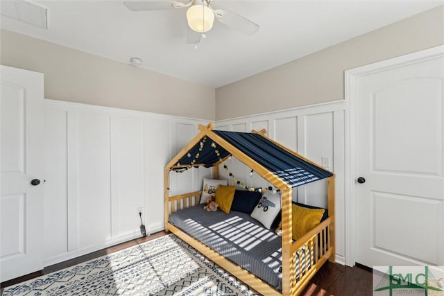 bedroom featuring a ceiling fan, a wainscoted wall, dark wood-style floors, visible vents, and a decorative wall