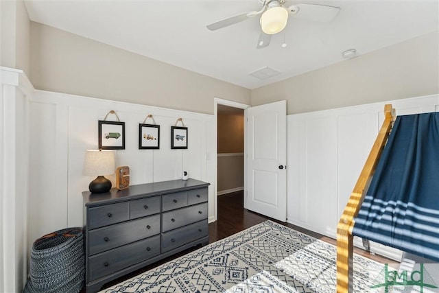 bedroom with a decorative wall, dark wood-type flooring, a ceiling fan, and a wainscoted wall