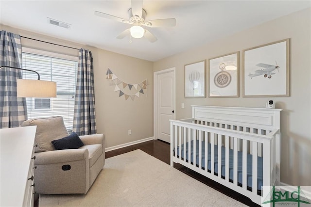 bedroom with wood finished floors, baseboards, visible vents, ceiling fan, and a nursery area