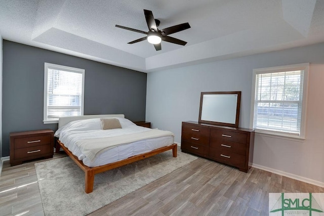 bedroom featuring a tray ceiling, multiple windows, baseboards, and light wood-style flooring