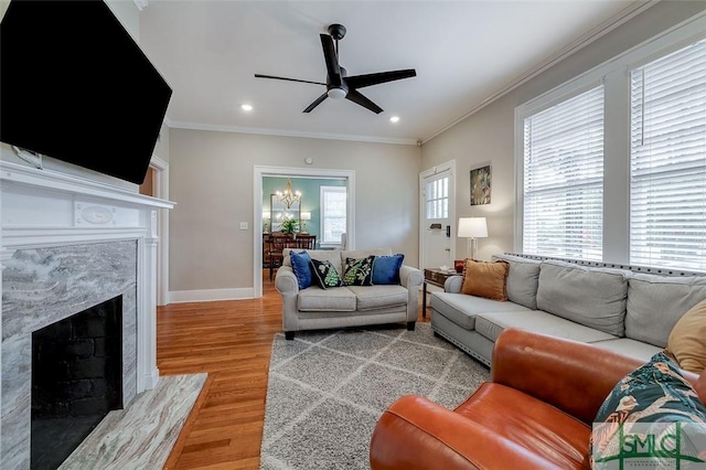 living room with crown molding, baseboards, a premium fireplace, ceiling fan with notable chandelier, and light wood-style floors