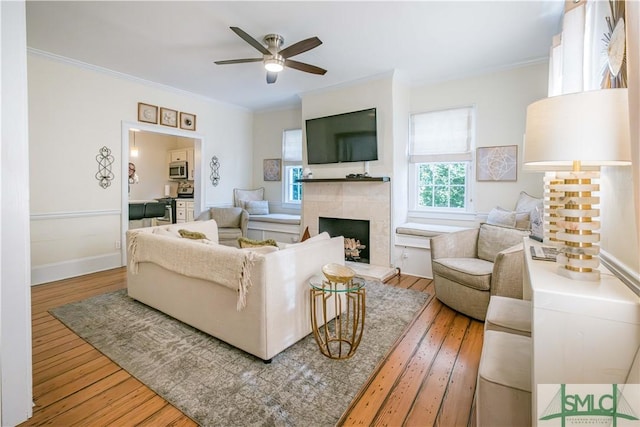 living area with baseboards, ceiling fan, a tile fireplace, hardwood / wood-style flooring, and crown molding