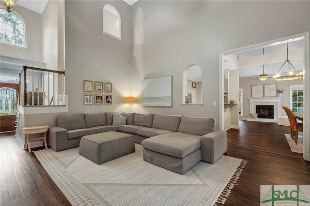 living room featuring a fireplace with raised hearth, ceiling fan with notable chandelier, dark wood-type flooring, and a towering ceiling