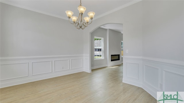 empty room featuring an inviting chandelier, arched walkways, a glass covered fireplace, crown molding, and light wood-type flooring