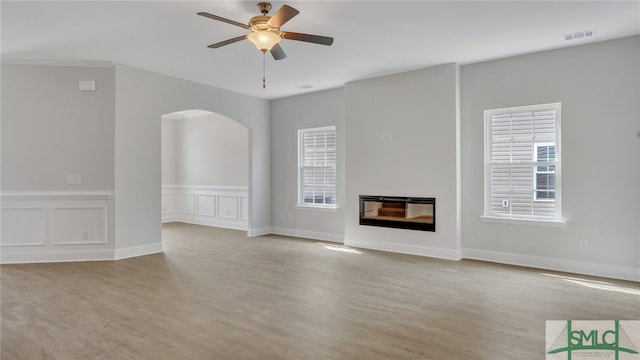 unfurnished living room featuring visible vents, light wood-style flooring, arched walkways, a glass covered fireplace, and a ceiling fan