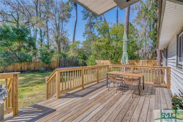 wooden terrace with outdoor dining area, a trampoline, a yard, and fence