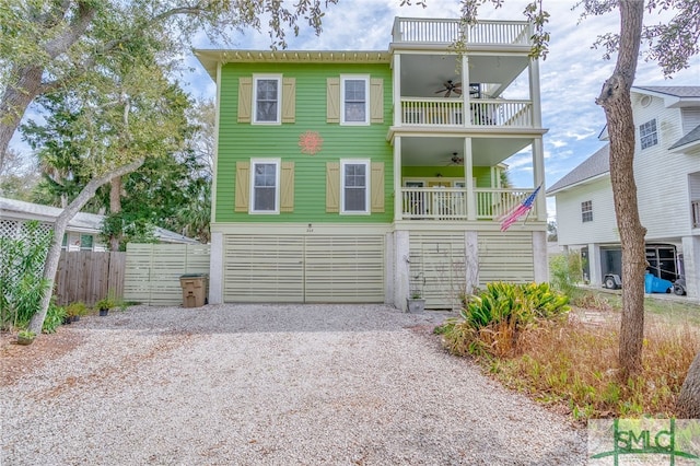 coastal home featuring fence, gravel driveway, an attached garage, a balcony, and ceiling fan