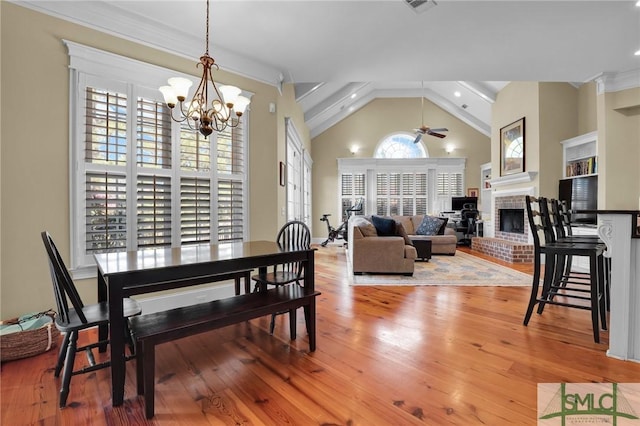 dining space with high vaulted ceiling, ceiling fan with notable chandelier, light wood-style floors, crown molding, and a brick fireplace