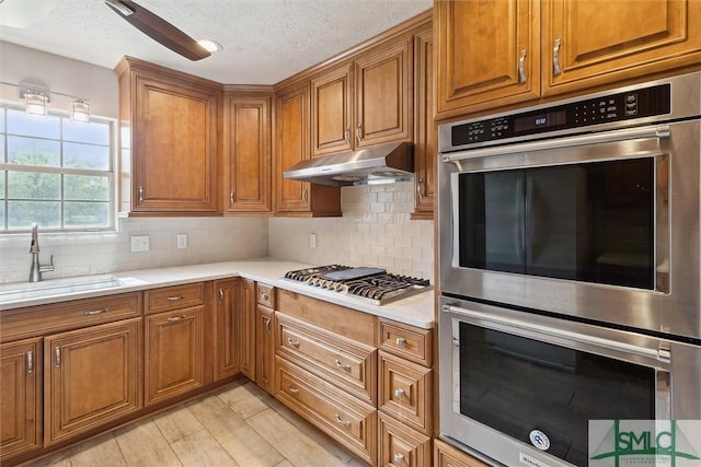 kitchen with a sink, brown cabinetry, under cabinet range hood, and stainless steel appliances