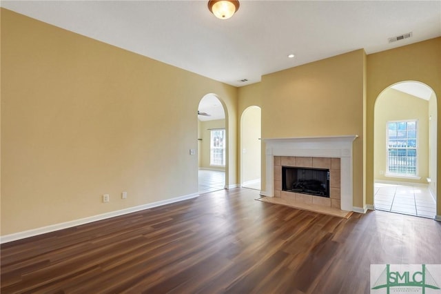 unfurnished living room featuring visible vents, baseboards, a tile fireplace, arched walkways, and dark wood-style flooring