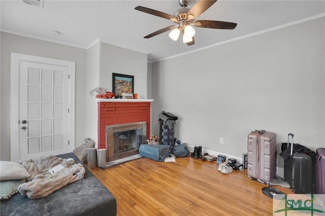 living room with a ceiling fan, baseboards, wood-type flooring, crown molding, and a brick fireplace