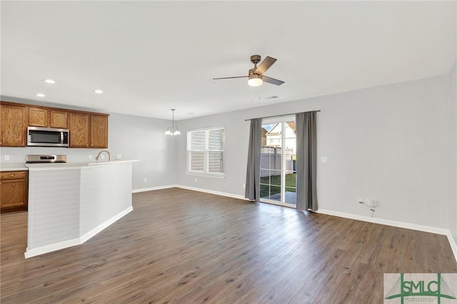 kitchen with ceiling fan with notable chandelier, stainless steel microwave, dark wood-style floors, brown cabinetry, and baseboards