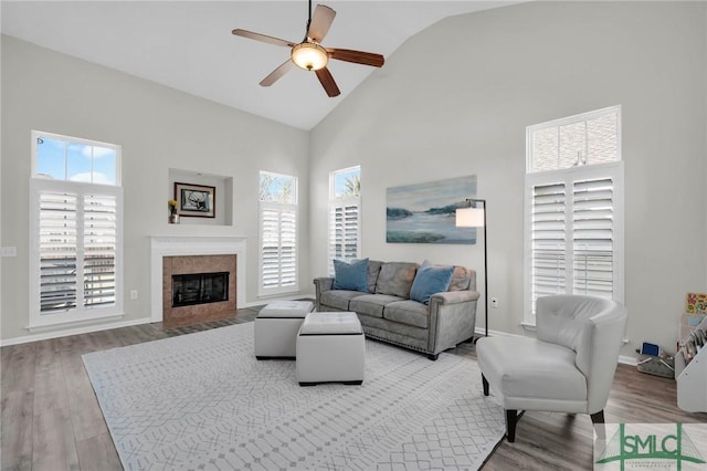 living area with high vaulted ceiling, a ceiling fan, a tiled fireplace, wood finished floors, and baseboards