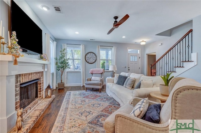 living room featuring wood finished floors, stairs, ceiling fan, a brick fireplace, and visible vents