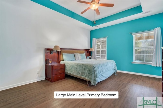 bedroom with ceiling fan, a tray ceiling, visible vents, baseboards, and wood finished floors