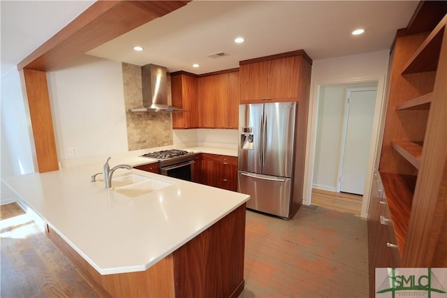 kitchen featuring brown cabinetry, stainless steel fridge, wall chimney range hood, a sink, and a peninsula