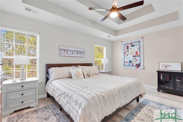 bedroom with light wood-type flooring, baseboards, a raised ceiling, ceiling fan, and visible vents