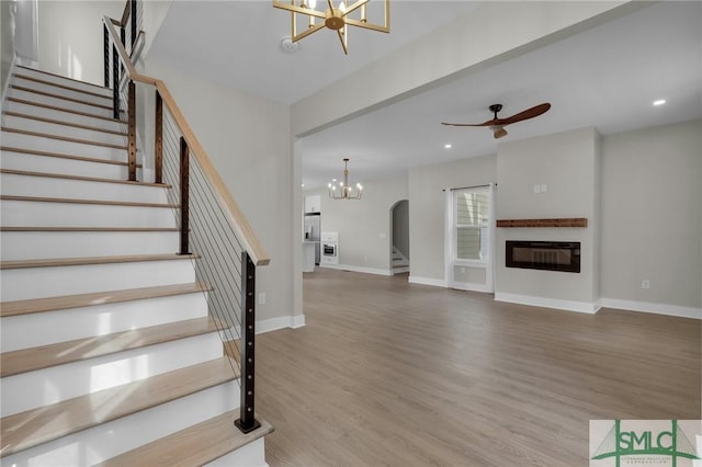 interior space with baseboards, a glass covered fireplace, ceiling fan with notable chandelier, wood finished floors, and stairway