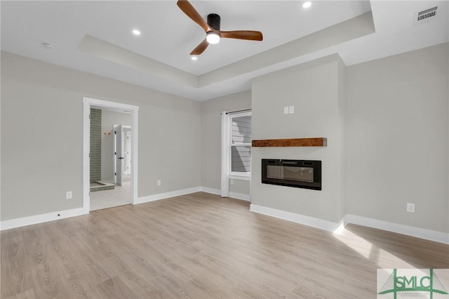 unfurnished living room featuring a tray ceiling, baseboards, a glass covered fireplace, ceiling fan, and light wood-type flooring