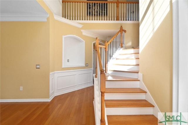staircase with a decorative wall, crown molding, wood finished floors, a high ceiling, and wainscoting