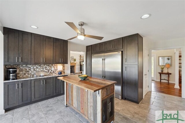 kitchen with built in refrigerator, butcher block countertops, decorative backsplash, a ceiling fan, and dark brown cabinetry