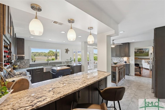 kitchen featuring decorative backsplash, visible vents, dark brown cabinetry, light stone counters, and hanging light fixtures