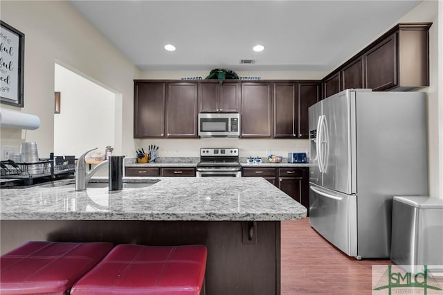 kitchen featuring appliances with stainless steel finishes, a peninsula, dark brown cabinetry, light stone countertops, and a breakfast bar area