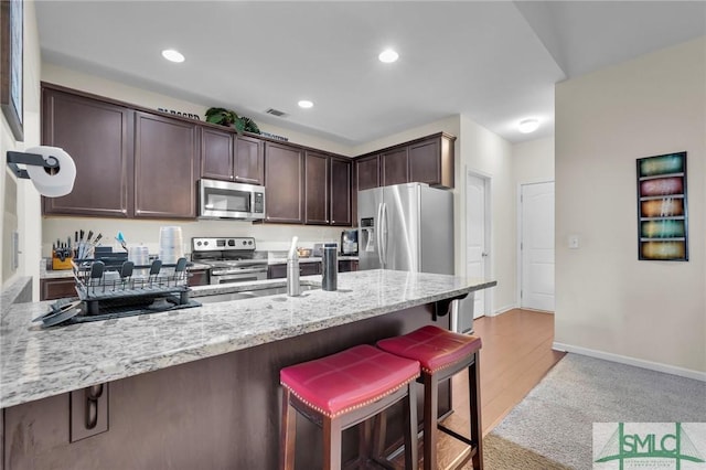 kitchen with dark brown cabinetry, a kitchen breakfast bar, a peninsula, light stone countertops, and appliances with stainless steel finishes