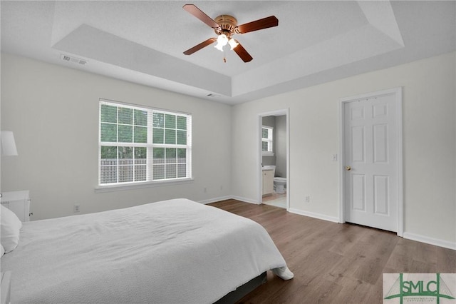 bedroom with wood finished floors, visible vents, a raised ceiling, connected bathroom, and baseboards