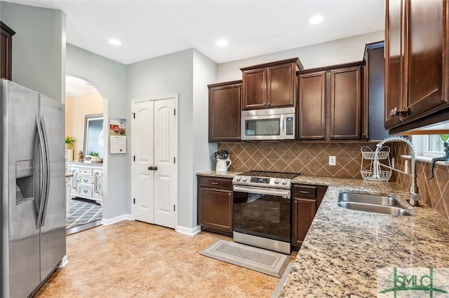 kitchen featuring dark brown cabinetry, arched walkways, light stone counters, a sink, and stainless steel appliances