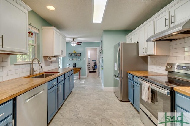kitchen featuring blue cabinets, a sink, under cabinet range hood, wooden counters, and appliances with stainless steel finishes