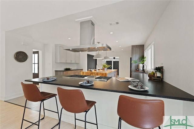 kitchen with island exhaust hood, a kitchen breakfast bar, stainless steel gas cooktop, visible vents, and dark countertops