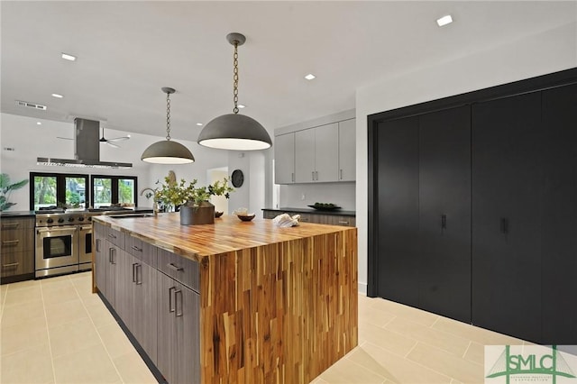 kitchen featuring wood counters, wall chimney exhaust hood, range with two ovens, a kitchen island with sink, and light tile patterned flooring