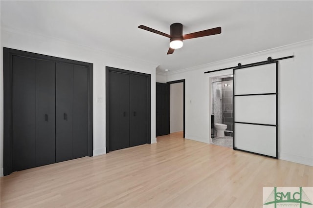unfurnished bedroom featuring light wood-style flooring, a barn door, ensuite bathroom, crown molding, and multiple closets
