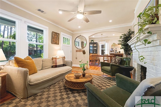 living room with wood finished floors, ceiling fan, arched walkways, ornamental molding, and a brick fireplace