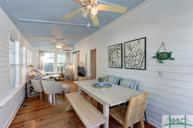 dining room with wood-type flooring, wooden walls, a wall mounted air conditioner, and ceiling fan