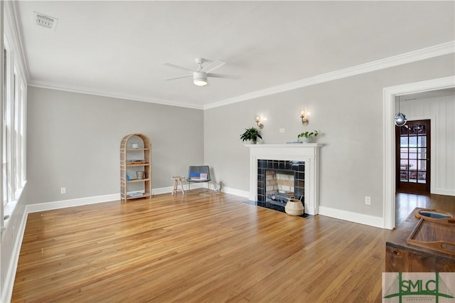 living area with a tiled fireplace, ceiling fan, visible vents, ornamental molding, and wood finished floors