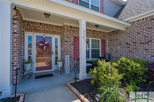 view of exterior entry featuring a shingled roof, covered porch, and brick siding