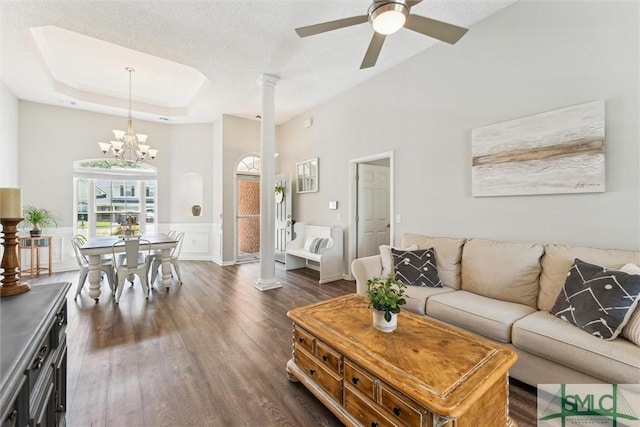 living area with dark wood-style floors, a raised ceiling, a wainscoted wall, ornate columns, and ceiling fan with notable chandelier
