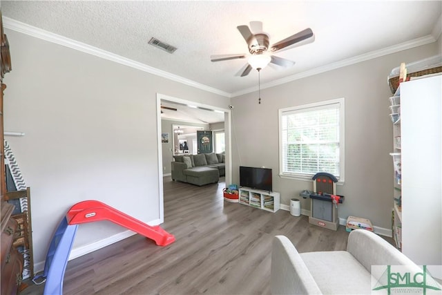recreation room with a ceiling fan, visible vents, wood finished floors, baseboards, and ornamental molding