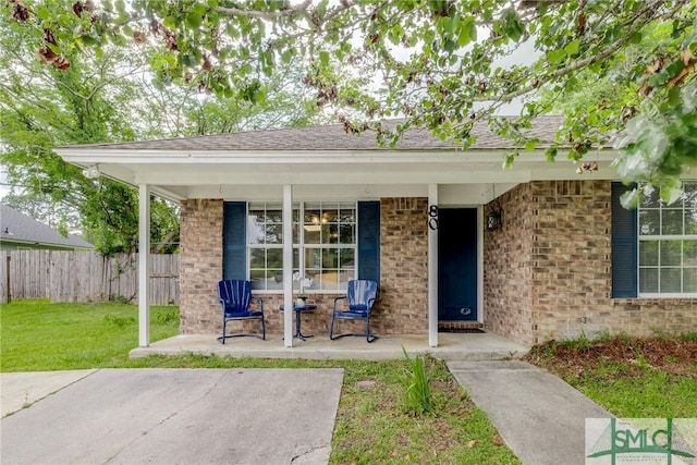 entrance to property with fence, roof with shingles, brick siding, a lawn, and covered porch