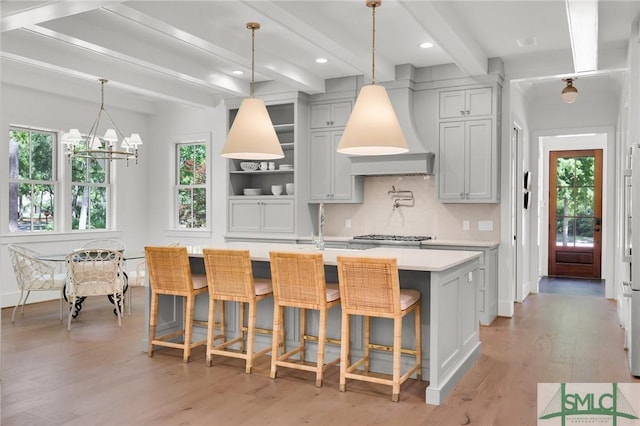 kitchen featuring beam ceiling, light wood finished floors, light countertops, custom range hood, and a center island with sink