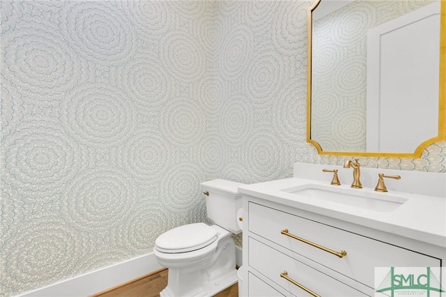 bathroom with toilet, wallpapered walls, wood finished floors, and vanity