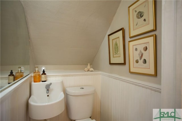 half bath featuring toilet, a textured ceiling, vaulted ceiling, and wainscoting
