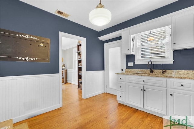 kitchen with a sink, a wainscoted wall, white cabinets, light wood finished floors, and light stone counters