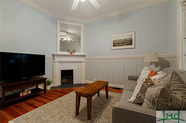 living room featuring a ceiling fan, wood finished floors, crown molding, a tile fireplace, and wainscoting