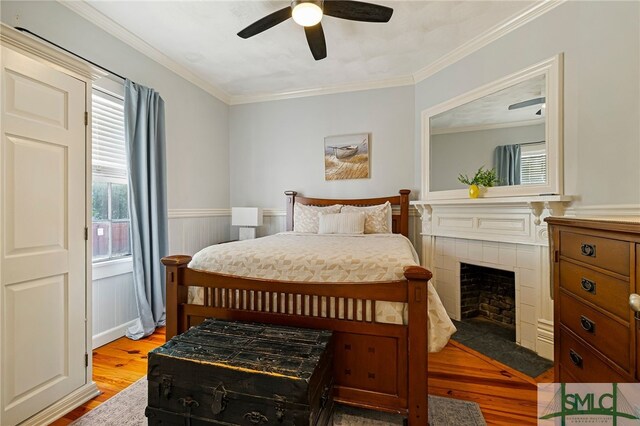 bedroom with light wood-style floors, ornamental molding, a tile fireplace, wainscoting, and a ceiling fan