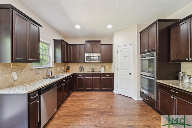 kitchen with stainless steel appliances, a sink, dark brown cabinetry, dark wood-style flooring, and light stone countertops